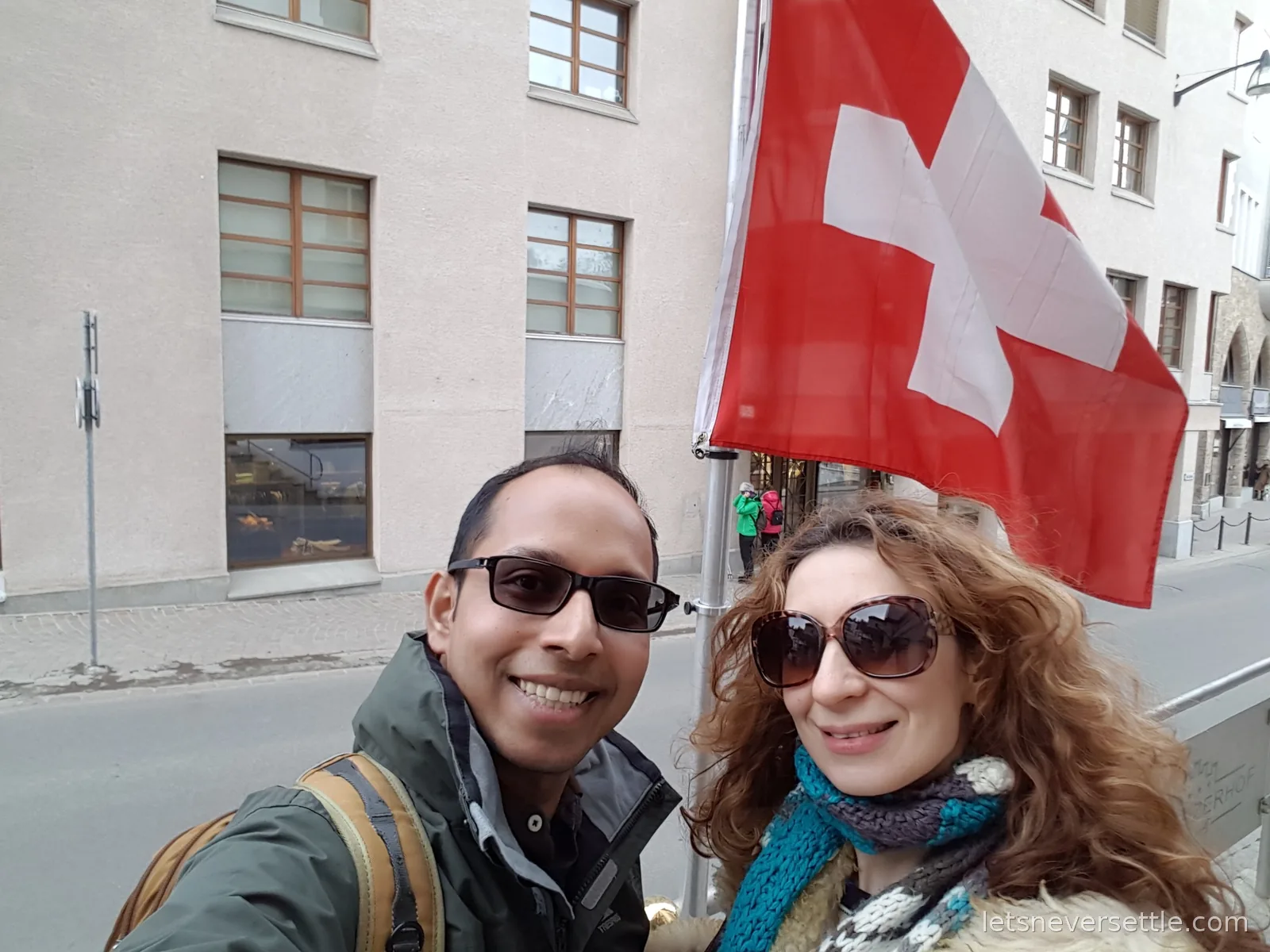 Inga and Dileep taking a selfie in sunglasses on a St. Moritz street with a large red and white Swiss flag hanging from a pole above them