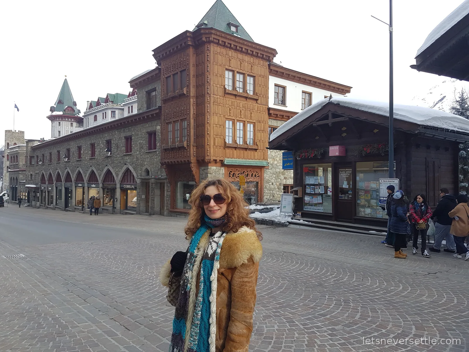 Inga on a wide cobbled St. Moritz street with an arcaded stone building, a timber clad corner house and a chalet style kiosk with snow on the roof