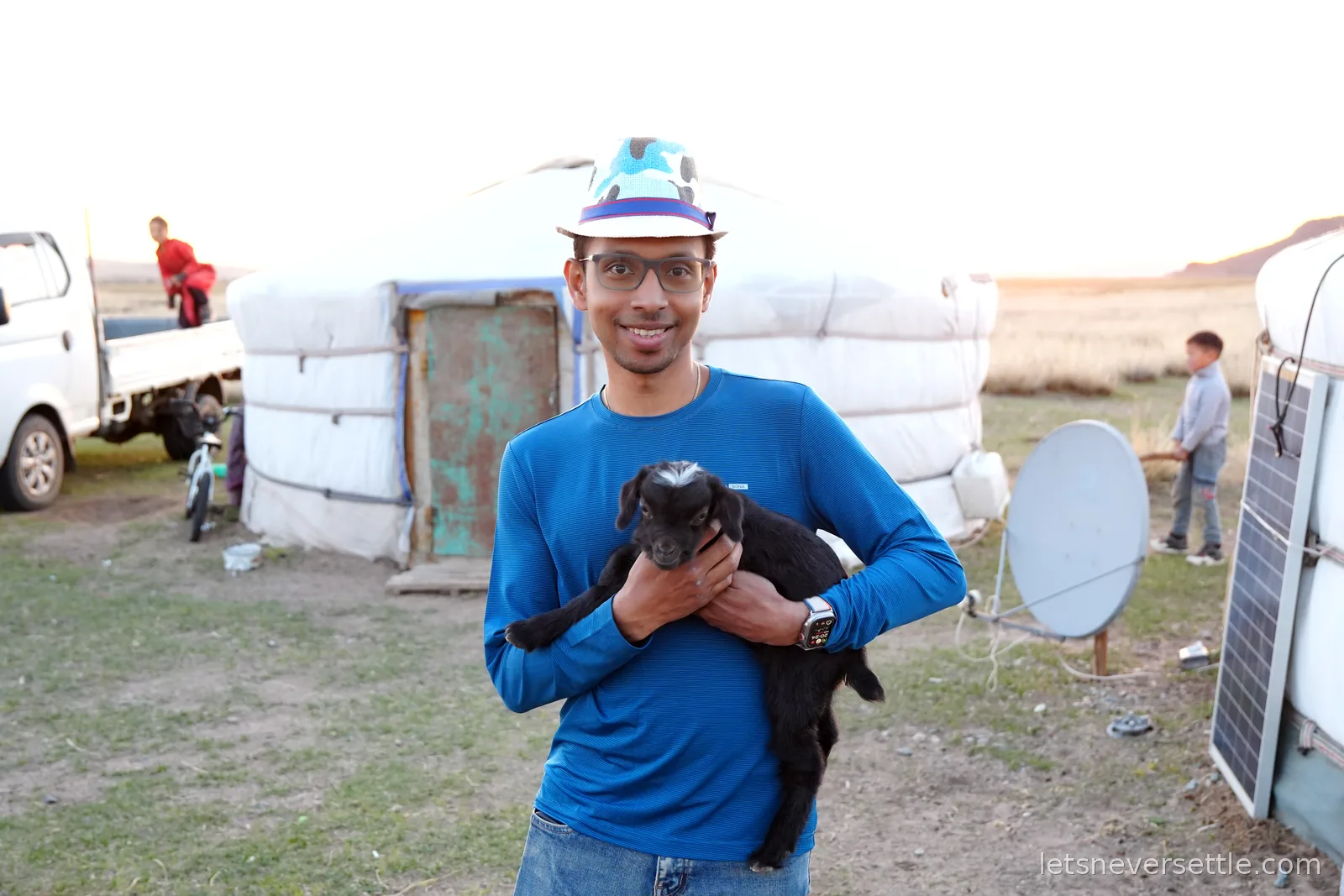 Dileep in a blue shirt and patterned hat holding a black baby goat in front of a ger, with a truck and satellite dish in the background
