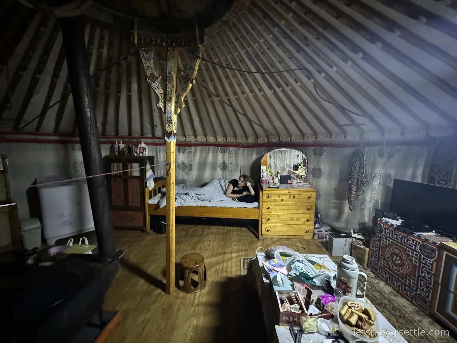 Inga lying on a wooden bed along the wall of the ger, with a dresser, mirror and low table with snacks and a thermos nearby