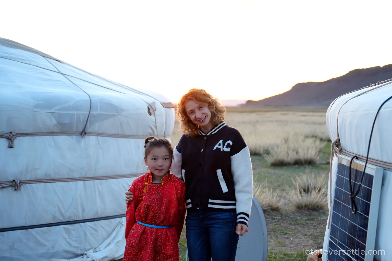 Inga with her arm around a young Mongolian girl in a red patterned deel, standing between two gers as the sun sets over the steppe
