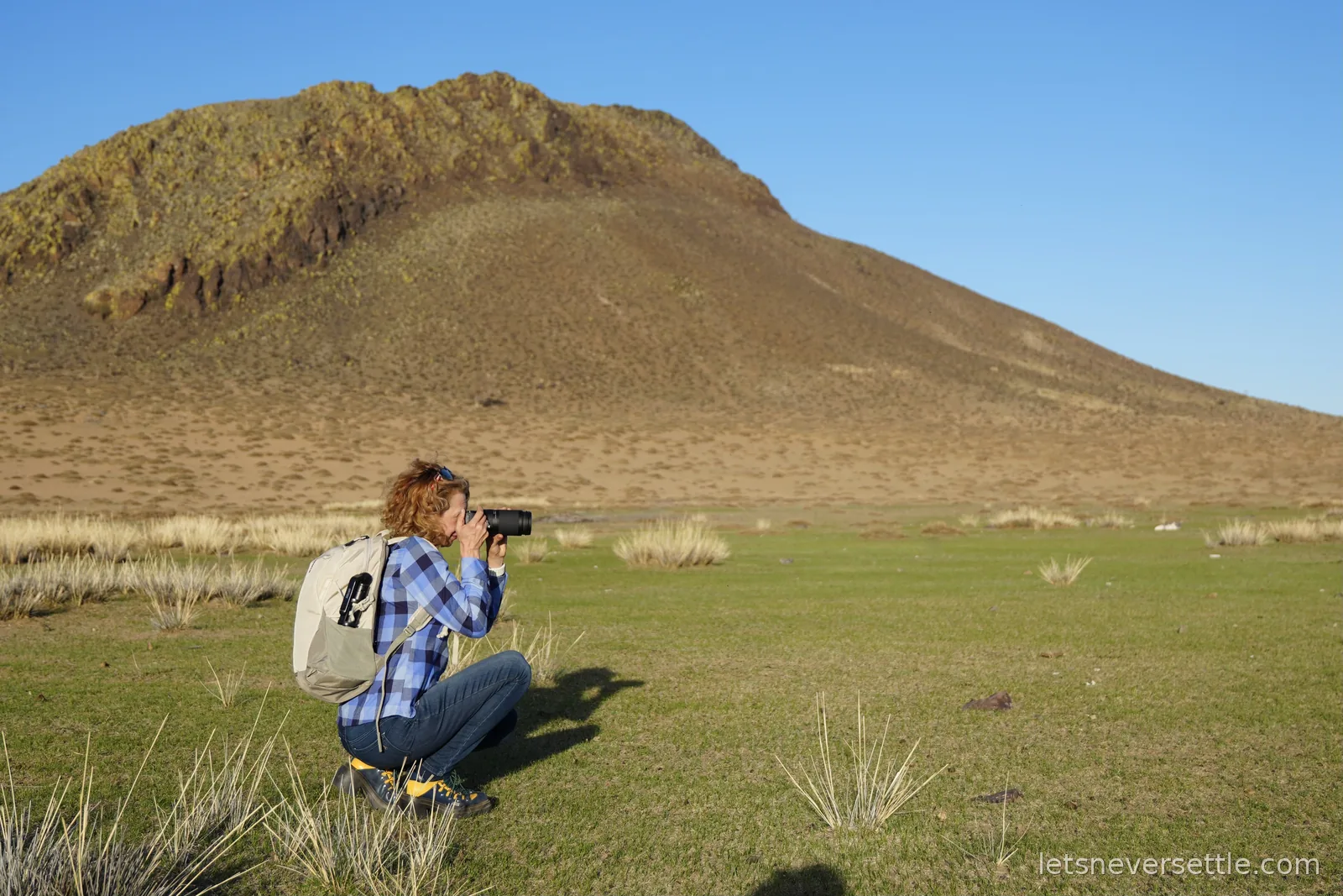 Inga crouching on green steppe grass with a camera raised, photographing a bare brown hill under a clear blue sky