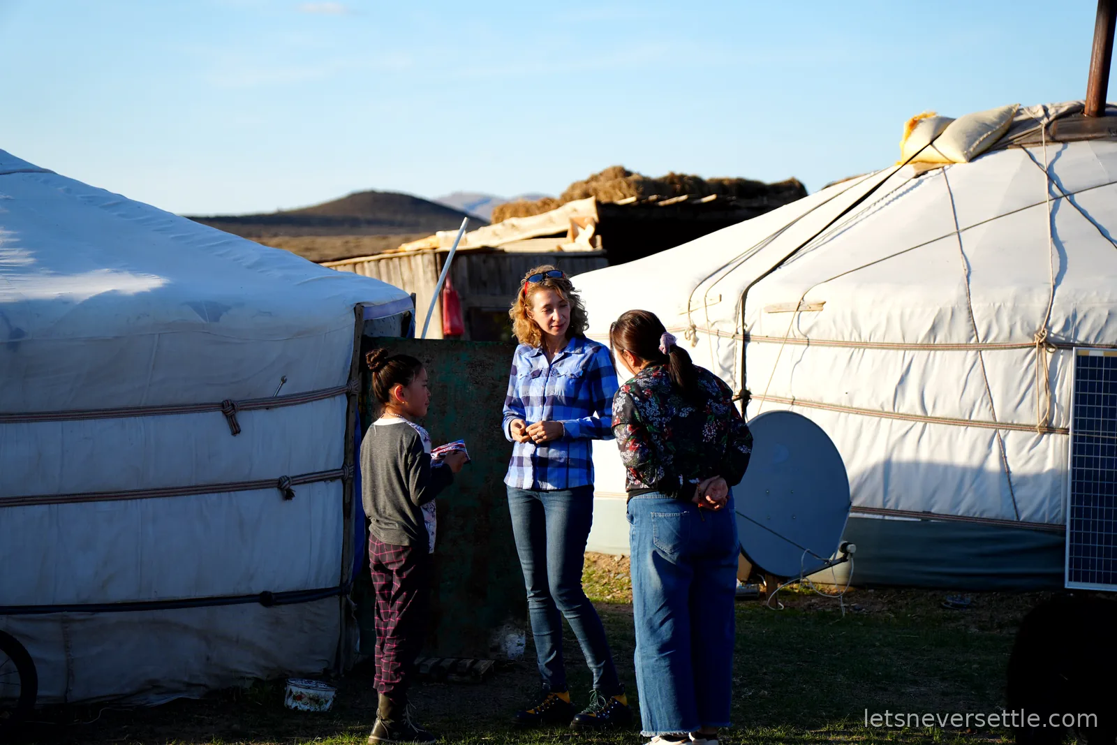 Inga chatting with a Mongolian woman and a young girl between two white gers, a satellite dish and solar panel beside them in evening light