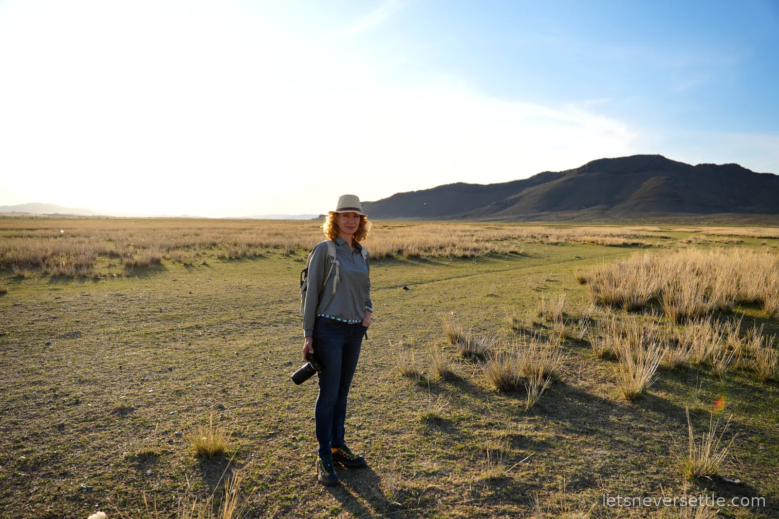 Inga standing in a sun hat with a camera in hand on open steppe grassland, low evening sun and dark hills on the horizon