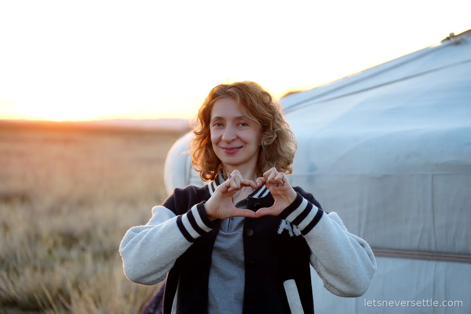 Inga making a heart shape with her hands in front of a ger, the sun setting into the grassland behind her