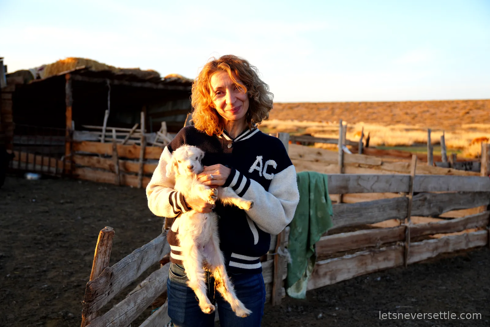 Inga in a varsity jacket cradling a white baby goat beside a wooden livestock pen, golden sunset light on the steppe behind
