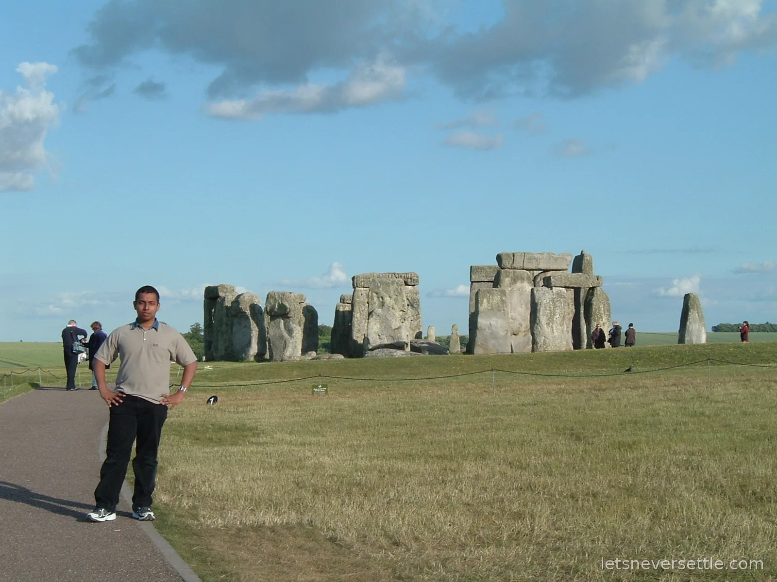 Dileep standing on the visitor path in front of the Stonehenge sarsen circle on a bright day, with the rope line and other visitors behind