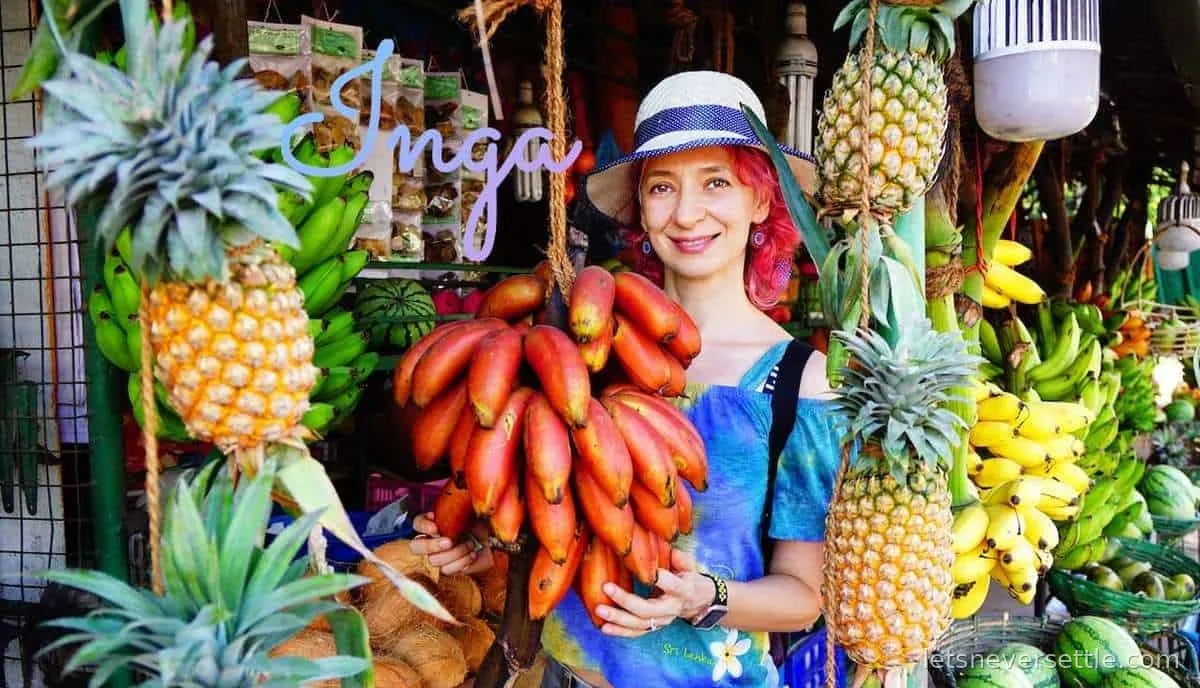Inga at a fruit market in Sri Lanka beside hanging bunches of red bananas and pineapples