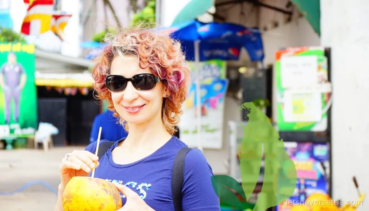Inga in sunglasses drinking from an opened orange king coconut through a straw at a shopfront in Sri Lanka