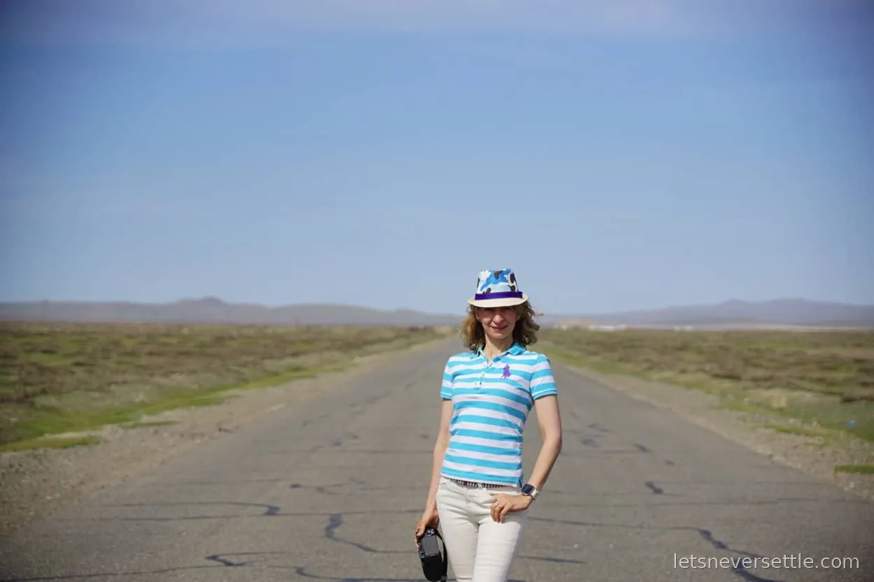 Inga standing on an empty tarmac road running straight across the Mongolian steppe under a wide blue sky
