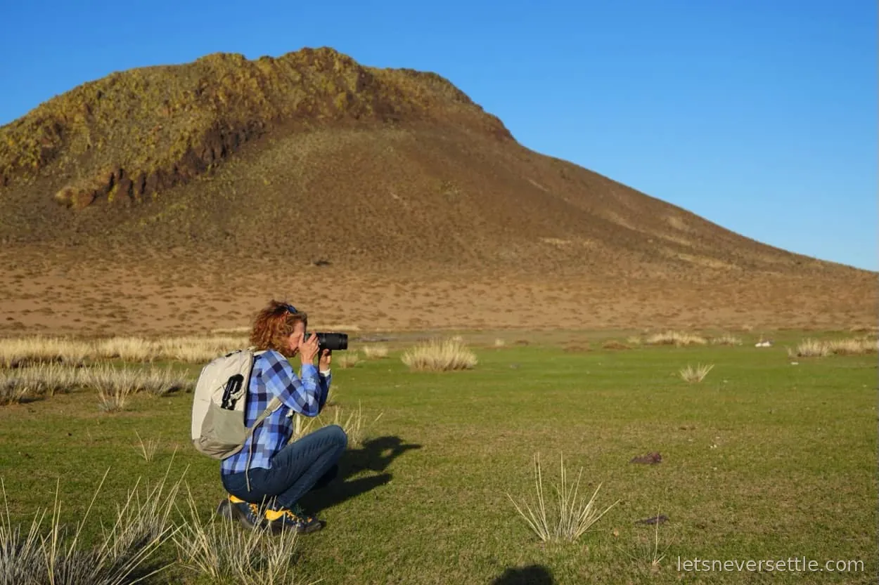 Inga crouching with a camera on the open steppe, a bare rocky hill rising behind her
