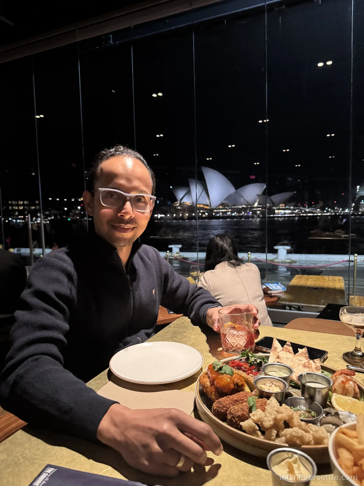 Dileep at a restaurant table with a share platter, the lit Opera House through the window behind