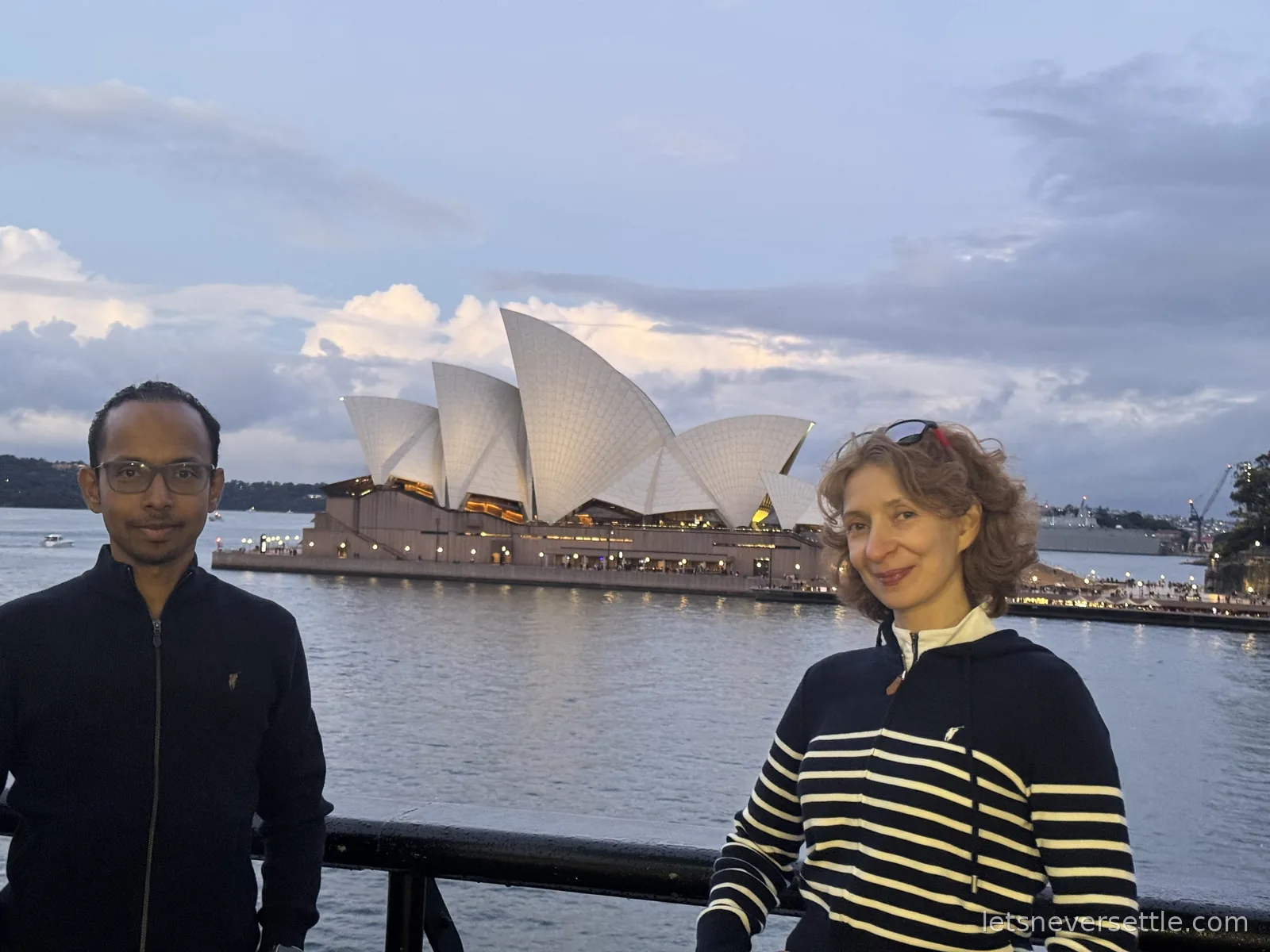Inga and Dileep either side of the Sydney Opera House seen lit across the water at dusk