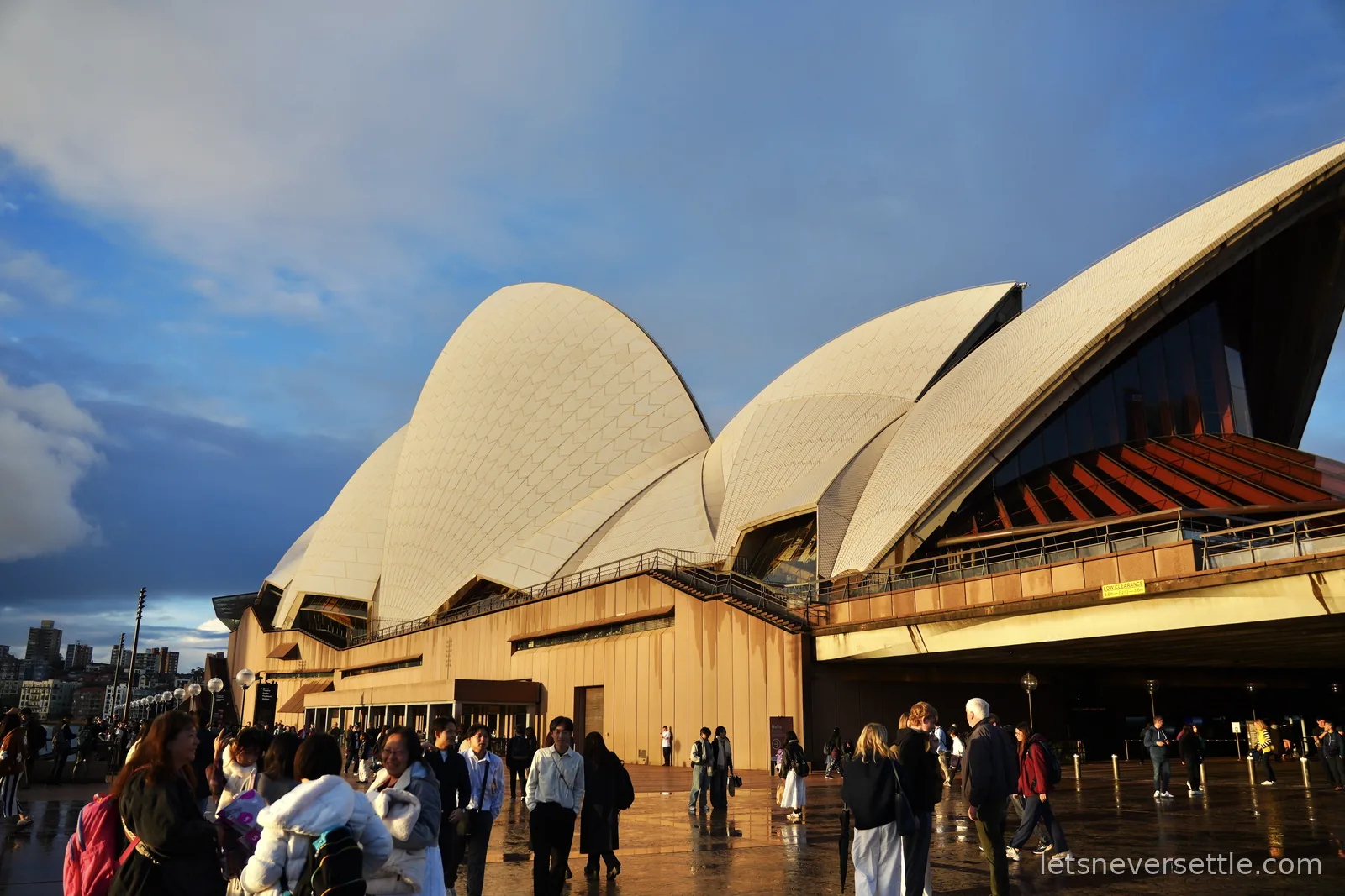 Sydney Opera House sails lit by low golden sun under a clearing sky, crowds on the wet forecourt
