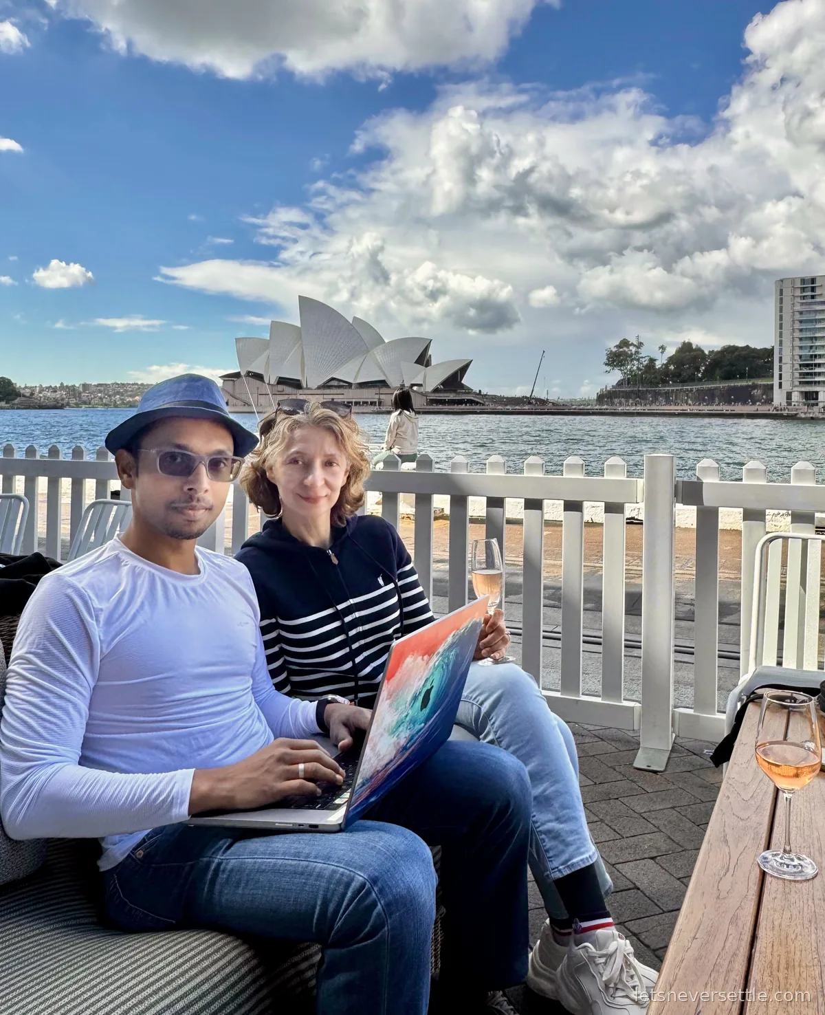 Inga and Dileep on a sofa by the Circular Quay waterfront with a laptop and glasses of rose wine, the Opera House across the harbour
