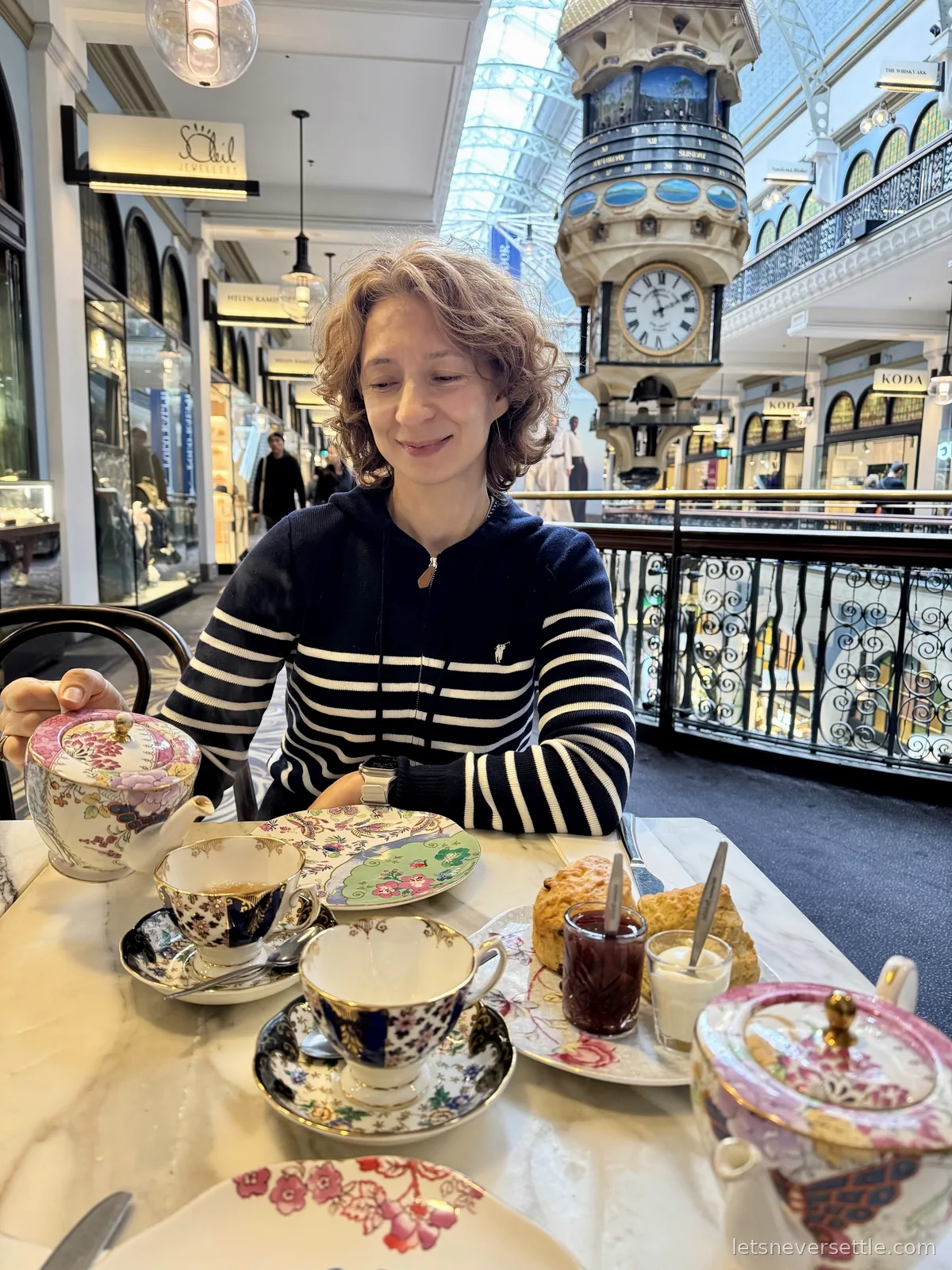 Inga pouring from a floral teapot at high tea in the QVB, scones with jam and cream on the table and the Great Australian Clock hanging behind