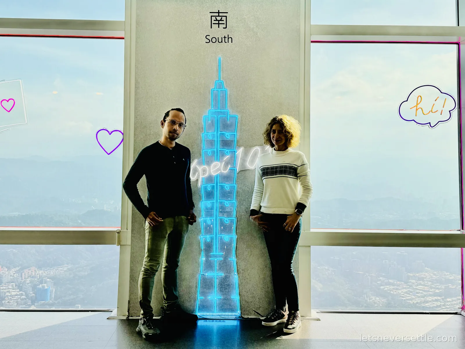 Inga and Dileep beside a neon outline of Taipei 101 at the tower's observatory, hazy mountains through the windows