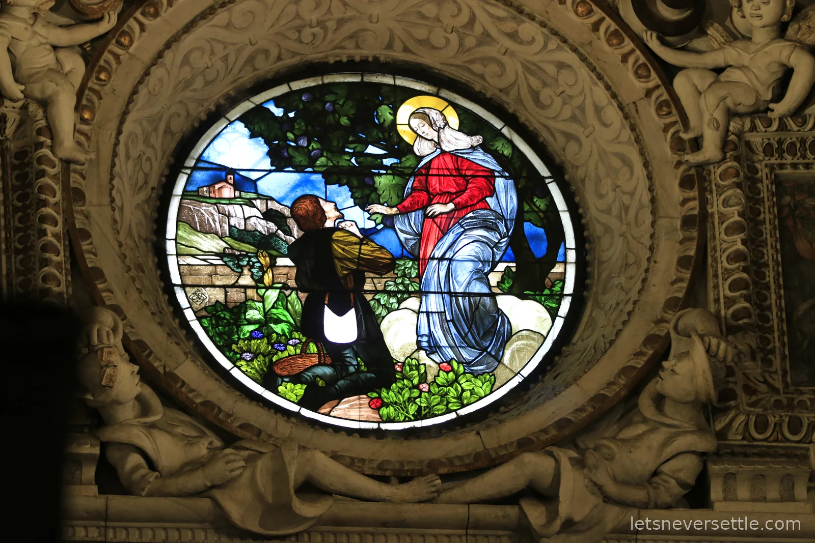 A circular stained glass window showing a robed woman appearing to a kneeling man on a mountain path, set in carved stone with cherubs at the corners
