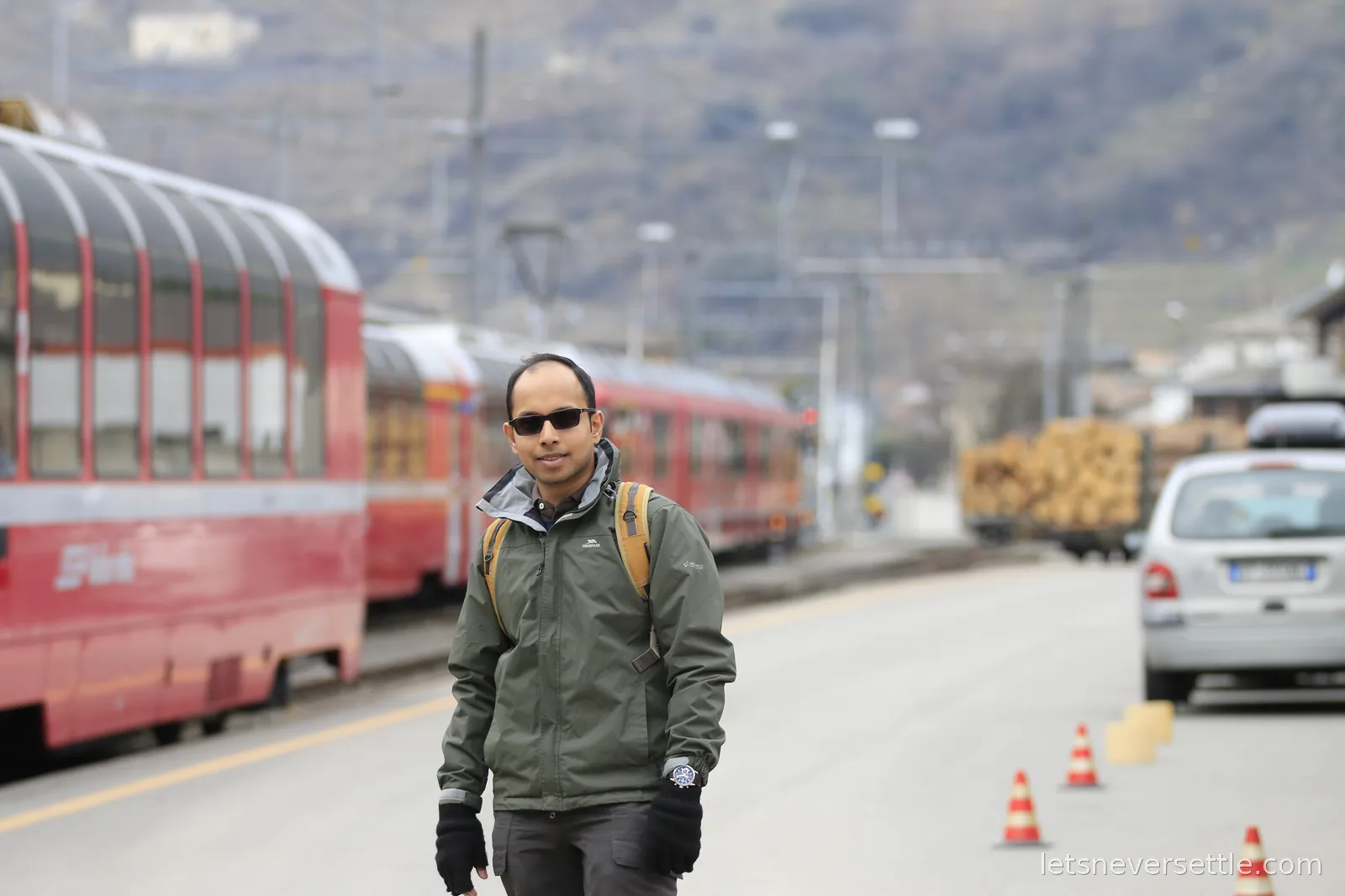 Dileep walking along the road beside a line of bright red Rhaetian Railway carriages with panoramic windows, mountains rising behind