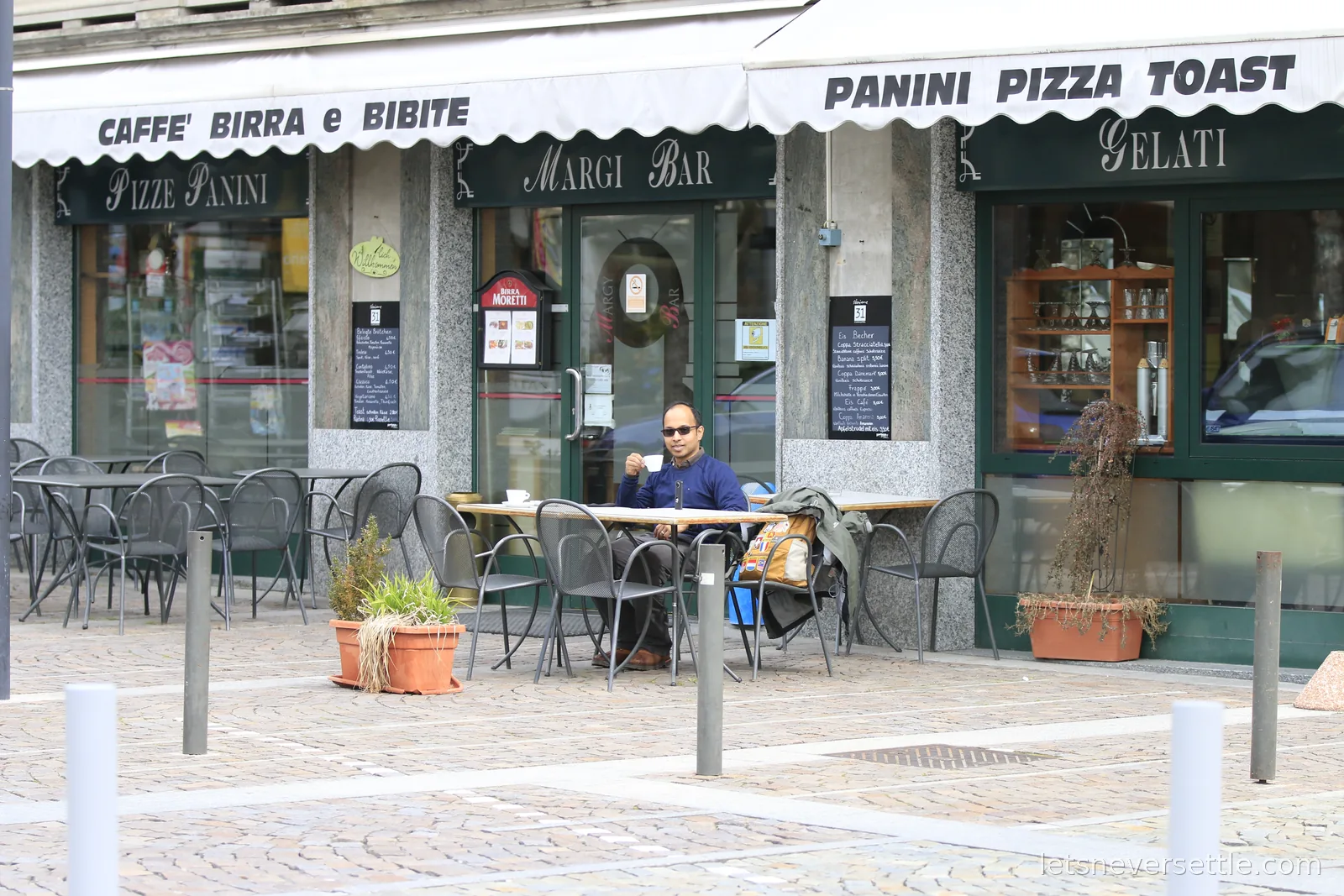 Dileep sitting alone at an outdoor table with a coffee outside a bar with green awnings lettered Caffe Birra e Bibite and Panini Pizza Toast