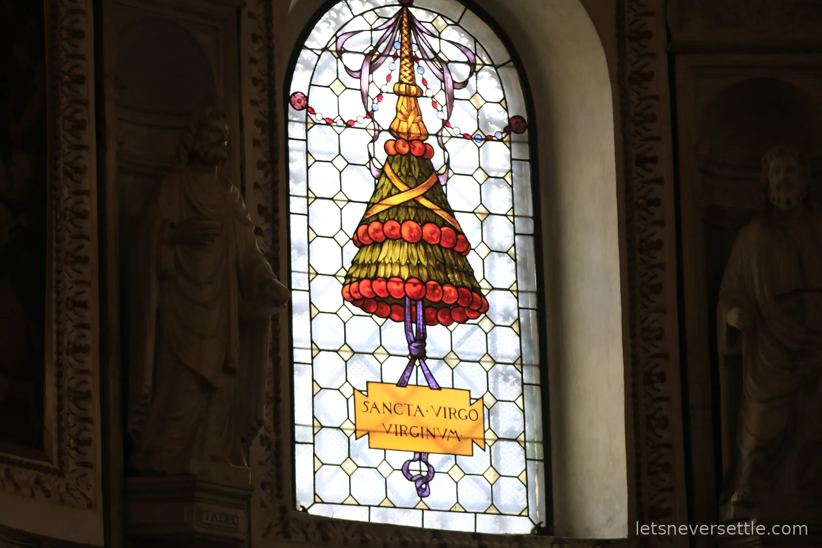 A tall stained glass window with a stylised gold and red design and a scroll lettered Sancta Virgo Virginum, flanked by stone statues