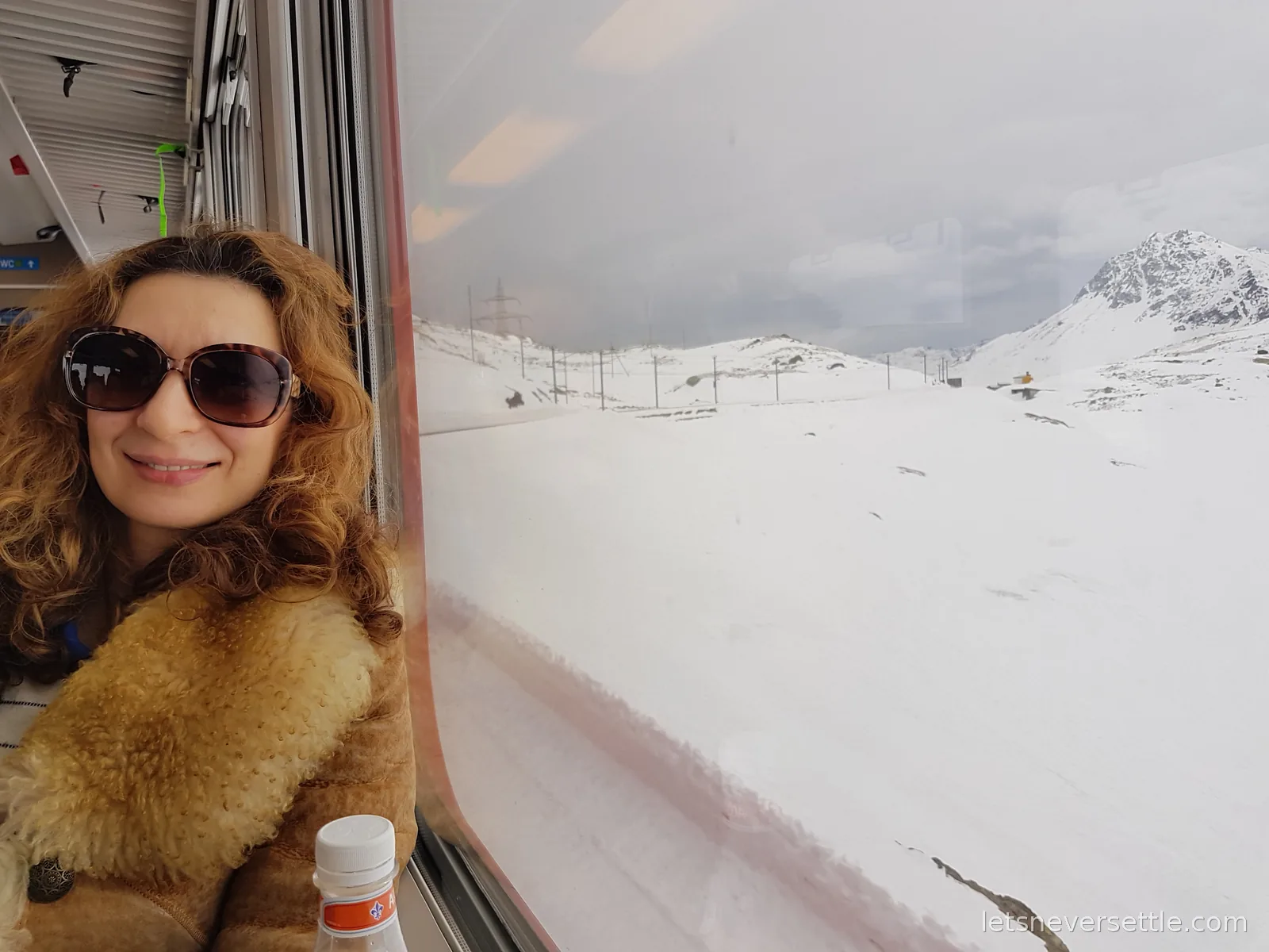 Inga smiling at a train window in sunglasses and a shearling coat, with a completely snow covered pass and telegraph poles outside