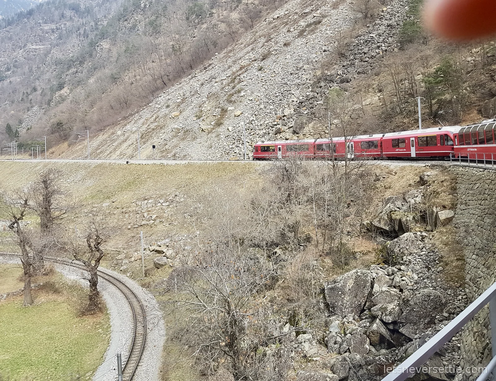The red train curving along a ledge on a steep scree slope, with the track it has already climbed looping back below through a field