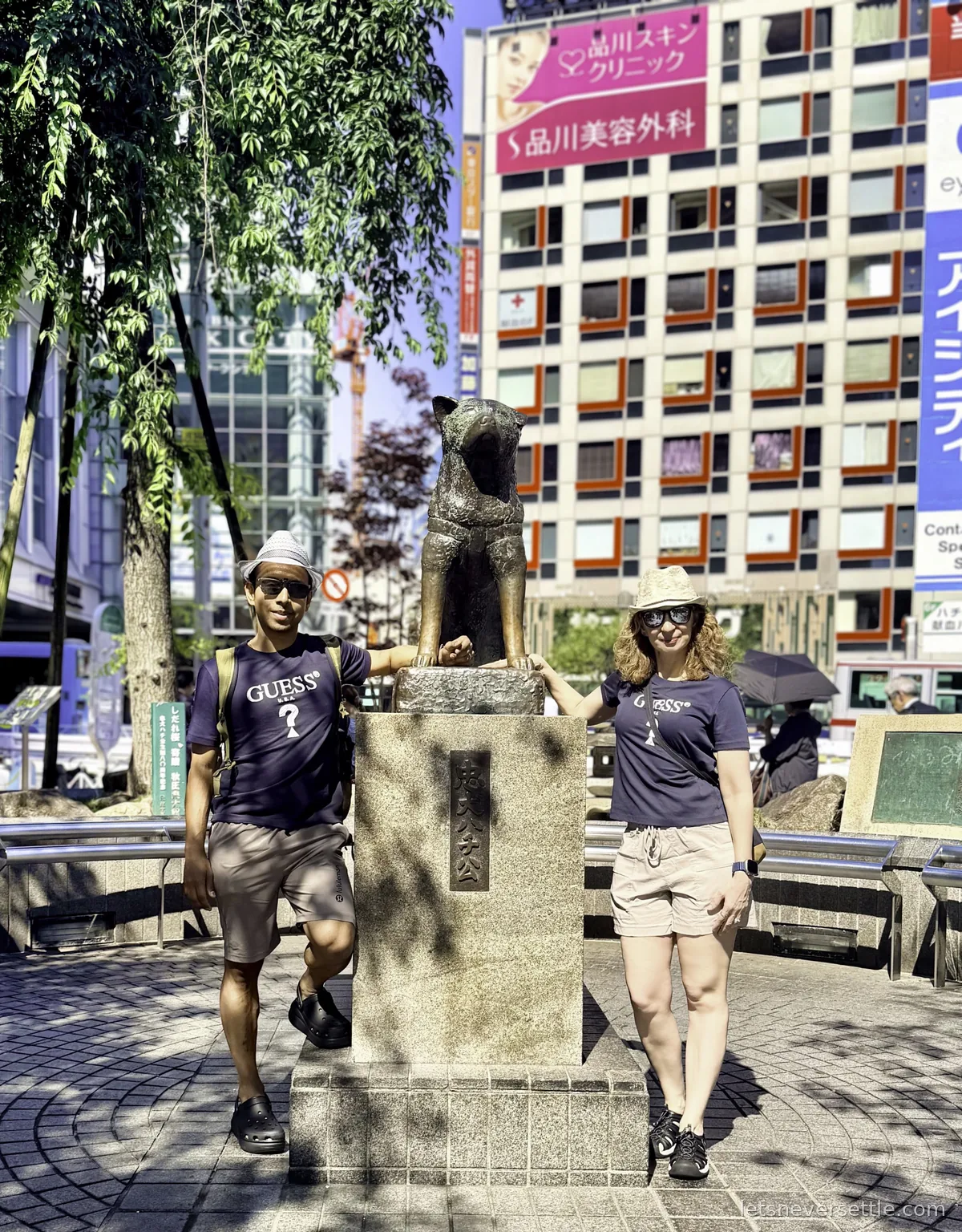 Dileep and Inga in sun hats and sunglasses standing either side of the bronze Hachiko dog statue outside Shibuya Station