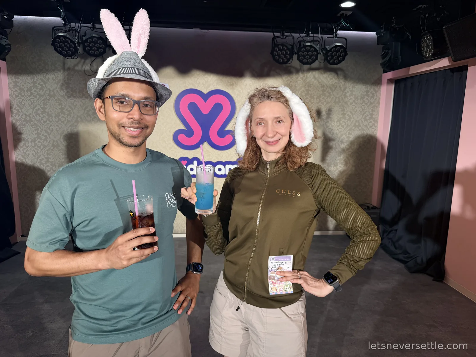 Dileep and Inga wearing pink bunny ears and holding drinks in front of the maidreamin heart logo on the cafe stage