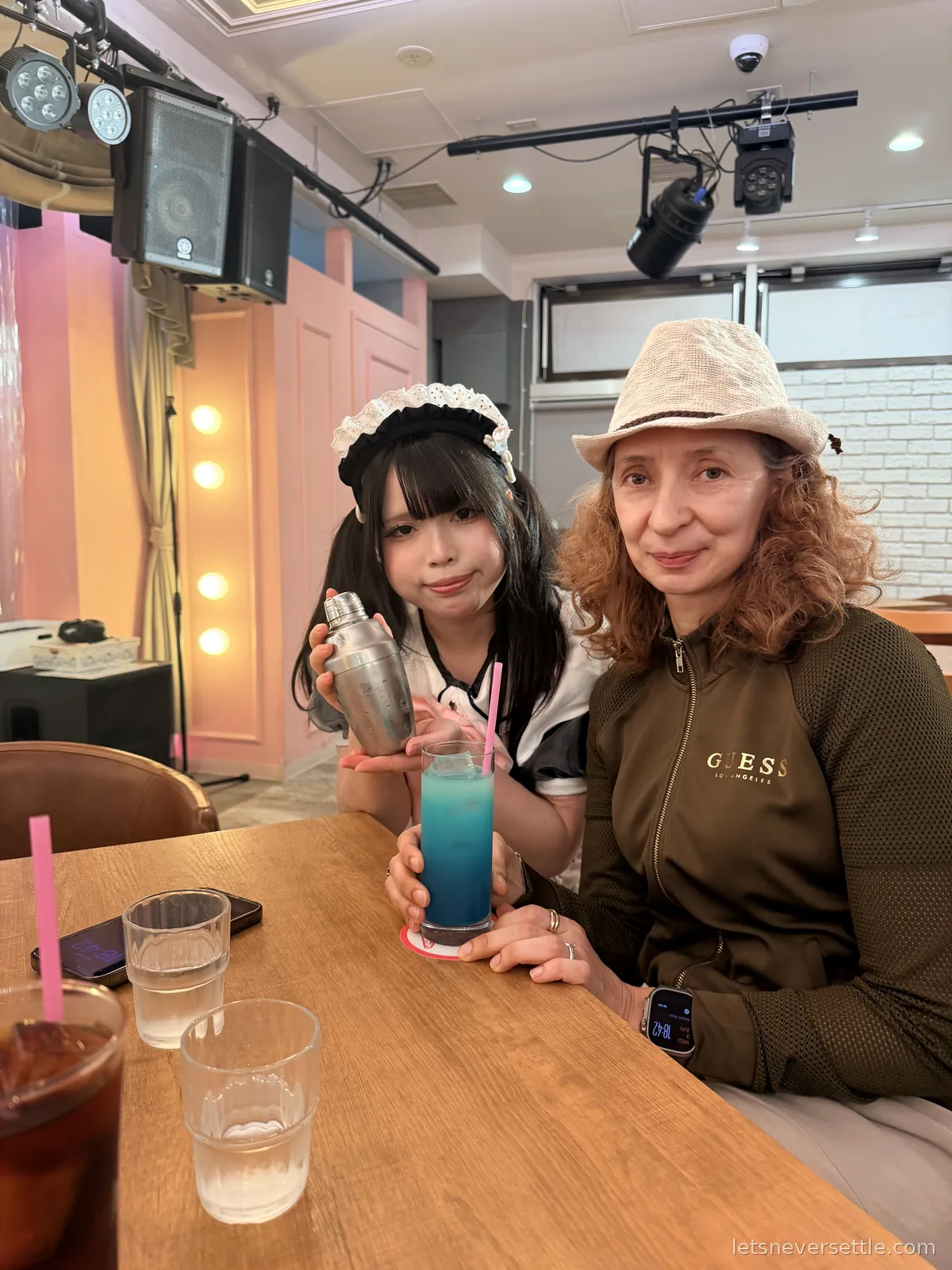 A maid in a black and white uniform holding a cocktail shaker beside Inga at a table, with a bright blue drink in front of them