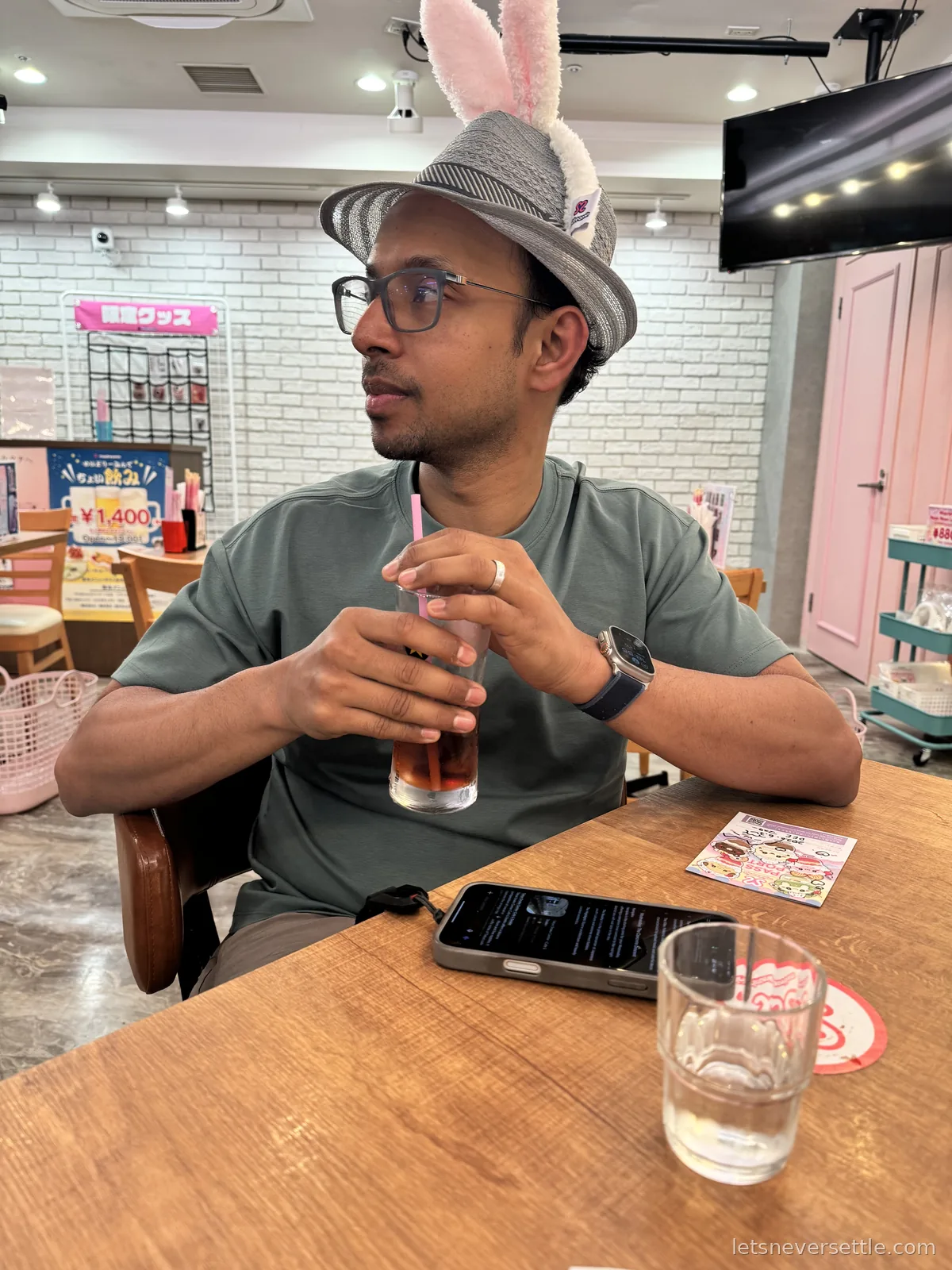 Dileep with pink bunny ears clipped to his straw hat, holding an iced tea at a wooden table in a maid cafe with pink fittings behind him
