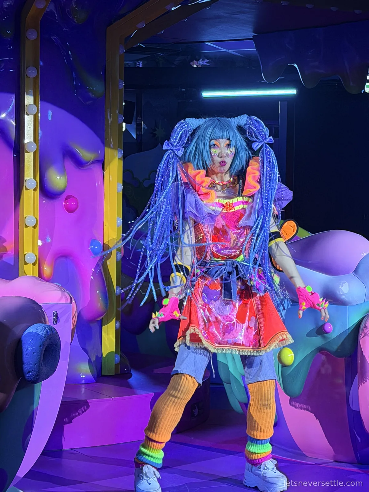 A performer with long blue braided hair, orange leg warmers and a pink and orange outfit posing beside a purple dessert sculpture
