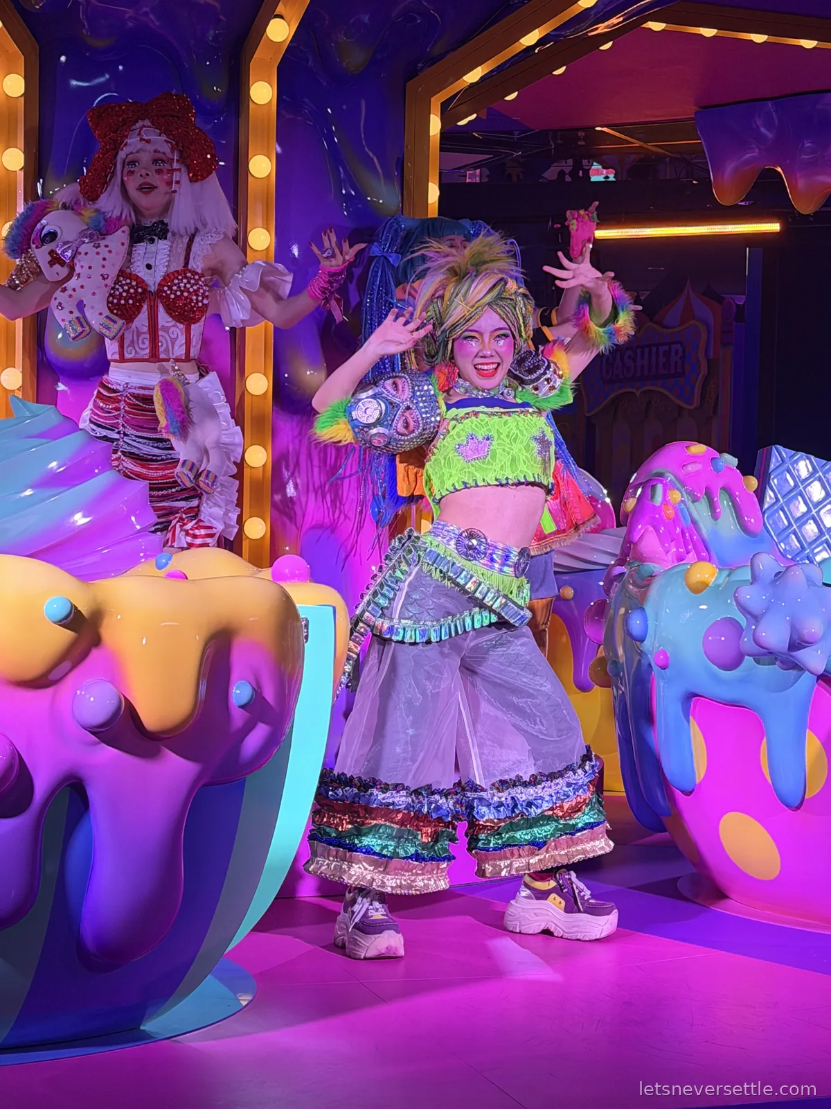 A performer in a neon green outfit dancing between giant melting dessert sculptures, with a second performer in a red wig on a lit-up stage behind