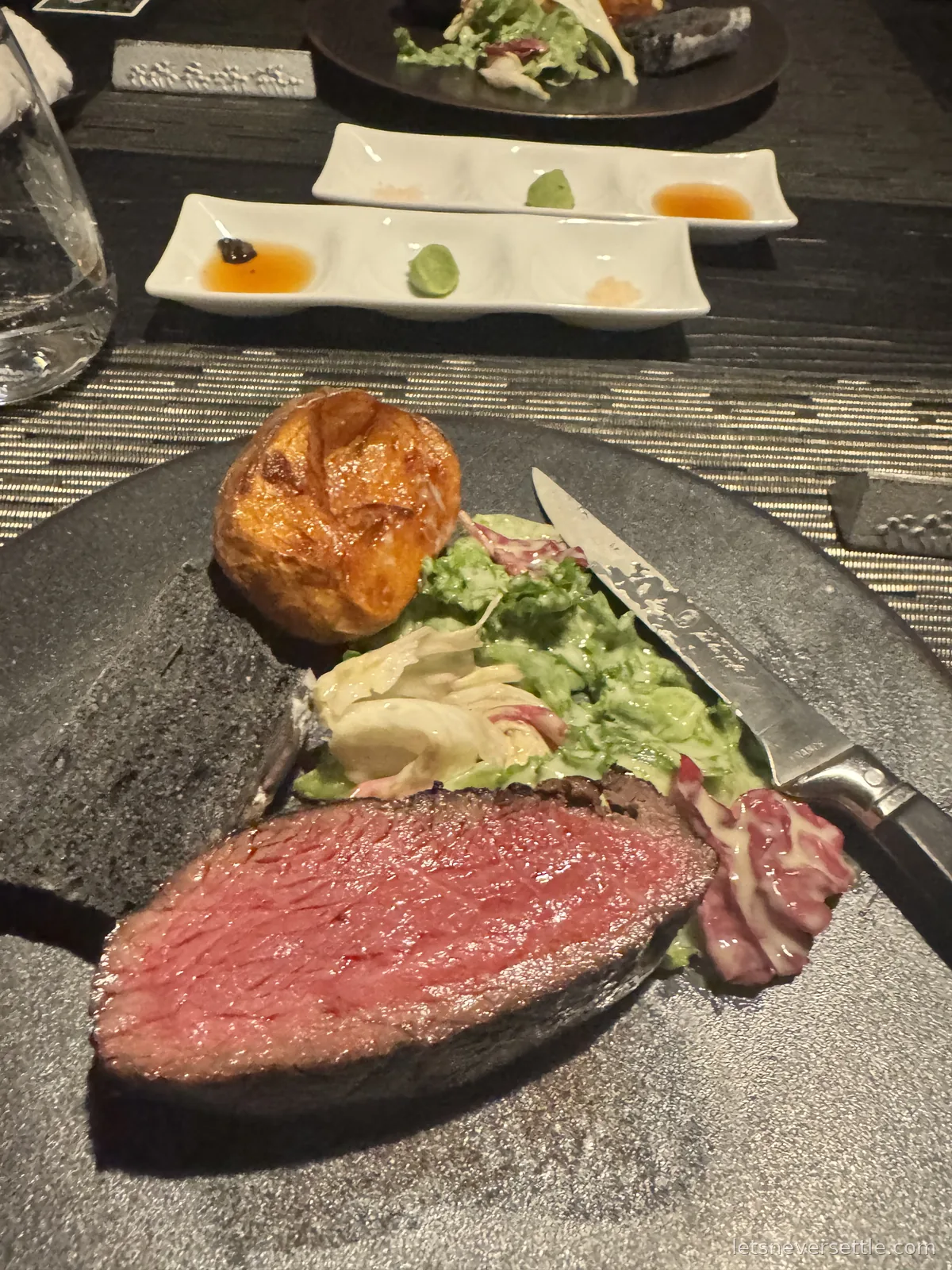 A thick slice of rare beef on a dark stone plate with a knife, dressed green salad and a roasted potato, with a tray of small condiments behind