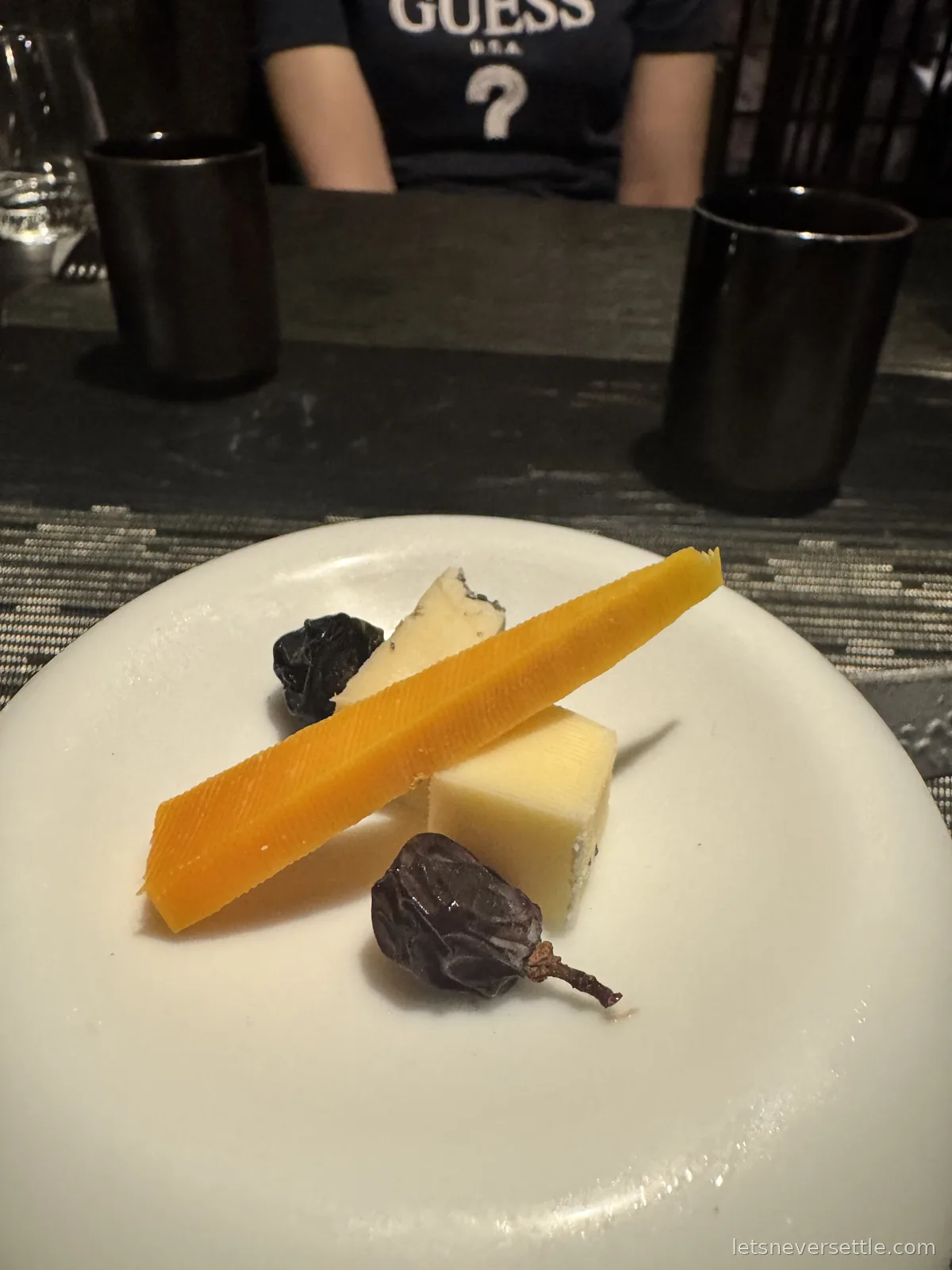 A white plate with three wedges of cheese and two dried fruits, with dark cups and a woven table mat behind