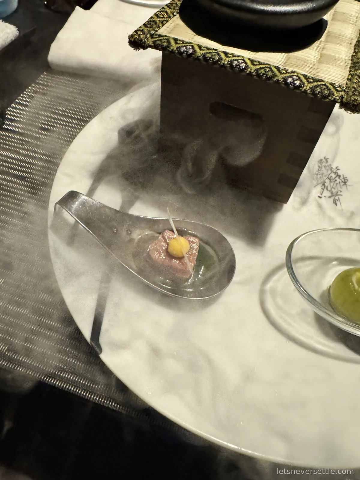 A single bite served in a glass spoon on a white plate surrounded by dry ice smoke, beside a small wooden box with a lidded pot on top