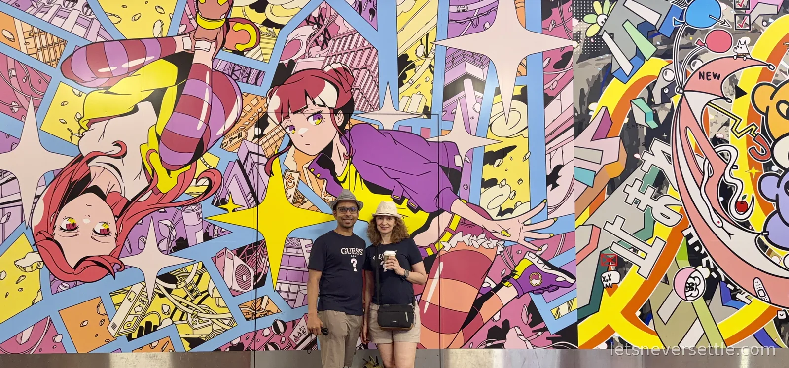 Dileep and Inga standing in front of a wall-sized manga mural in Shibuya covered in pink-haired characters, stars and city scenes