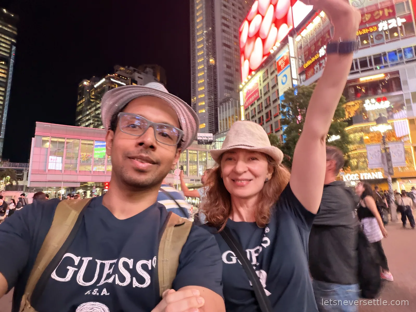 Night selfie of Dileep and Inga at Shibuya Crossing with Inga's arm raised, neon signs and crowds filling the background