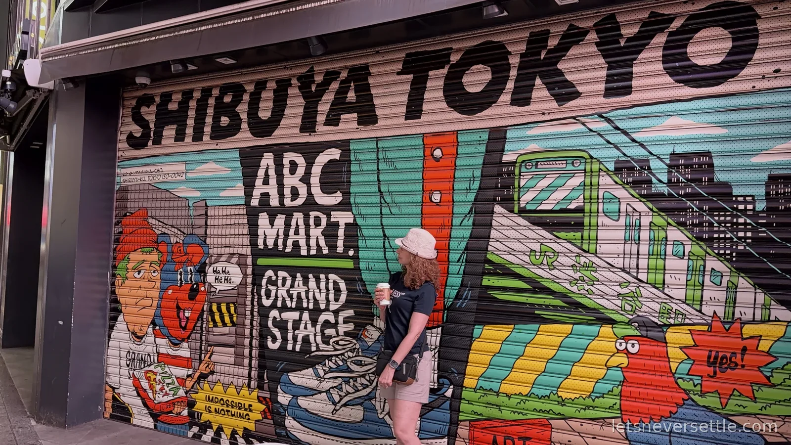 Inga holding a coffee in front of a painted ABC Mart shutter reading Shibuya Tokyo, covered in cartoon trainers, a JR train and a rooster