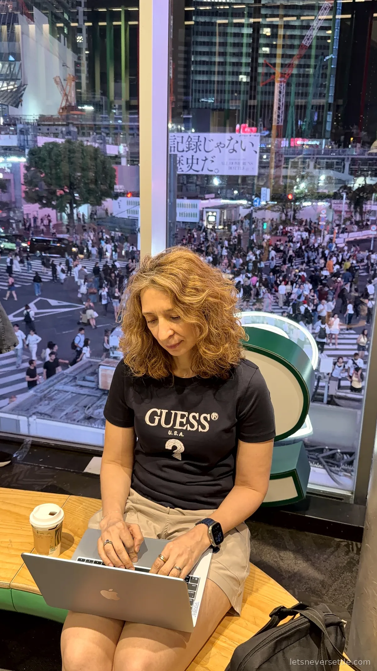 Inga working on a laptop at a window counter overlooking Shibuya Crossing at night, with a paper coffee cup beside her