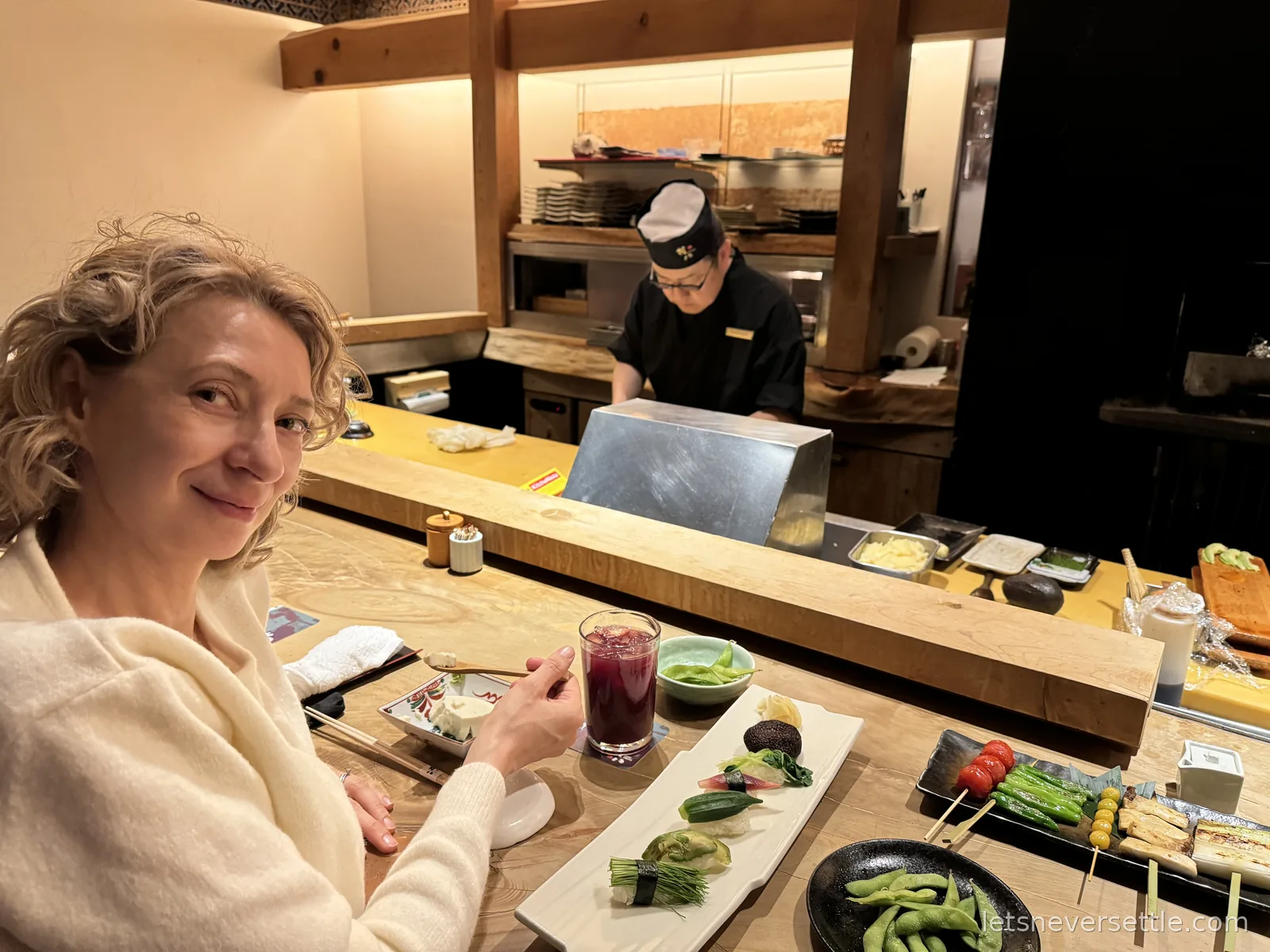 Inga at a wooden restaurant counter with plates of vegetable skewers and edamame while a chef works behind the bar