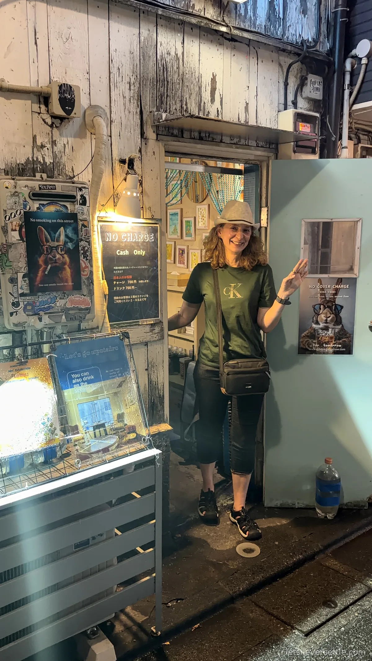Inga waving from the doorway of a tiny weathered bar in Golden Gai, Shinjuku, next to a sign reading No Charge, Cash Only