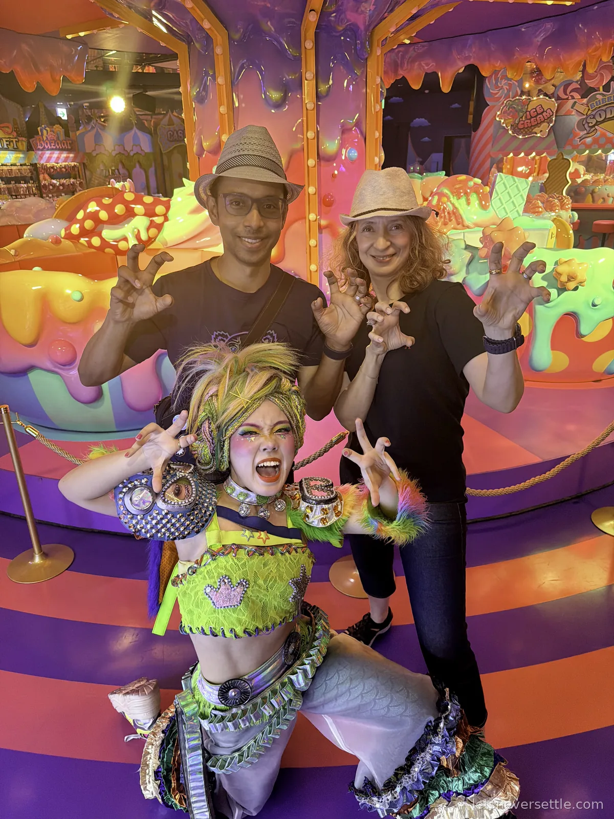 Inga and Dileep posing with clawed hands behind a performer in a neon monster costume in front of a candy-coloured carousel