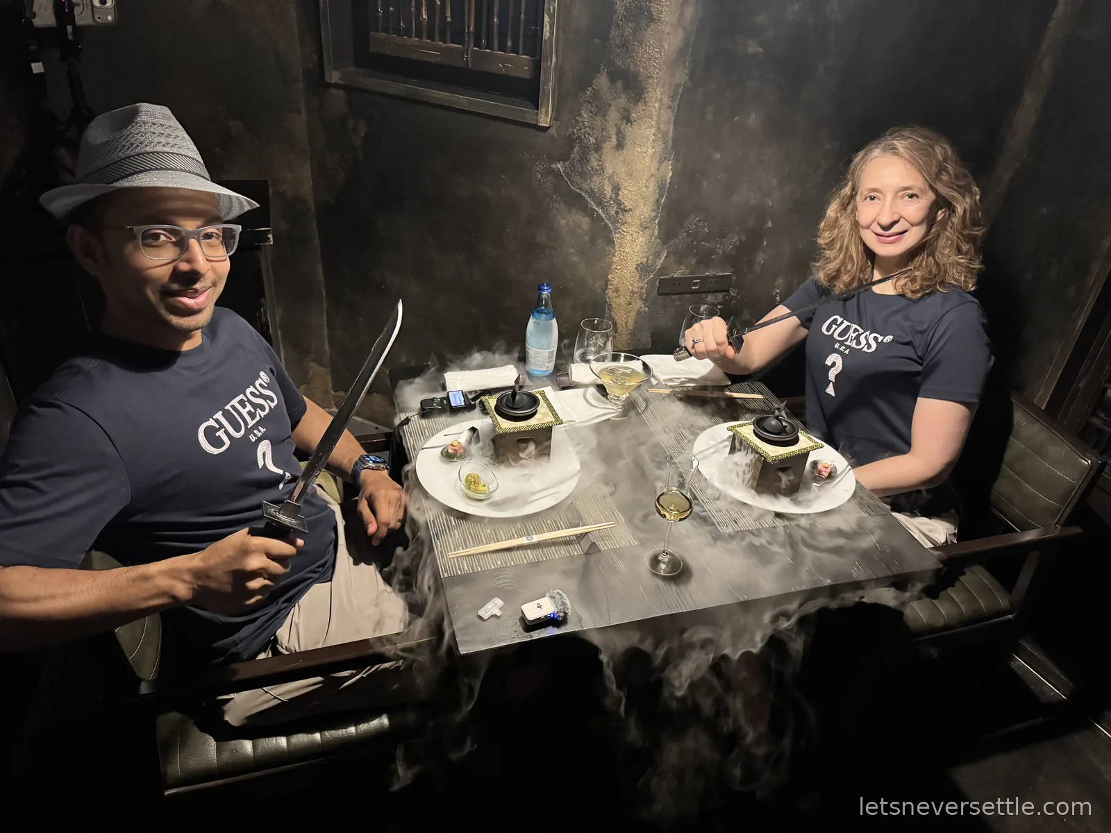 Dileep holding a katana beside a dinner table covered in dry ice smoke in a dimly lit ninja-themed restaurant, with Inga reaching across