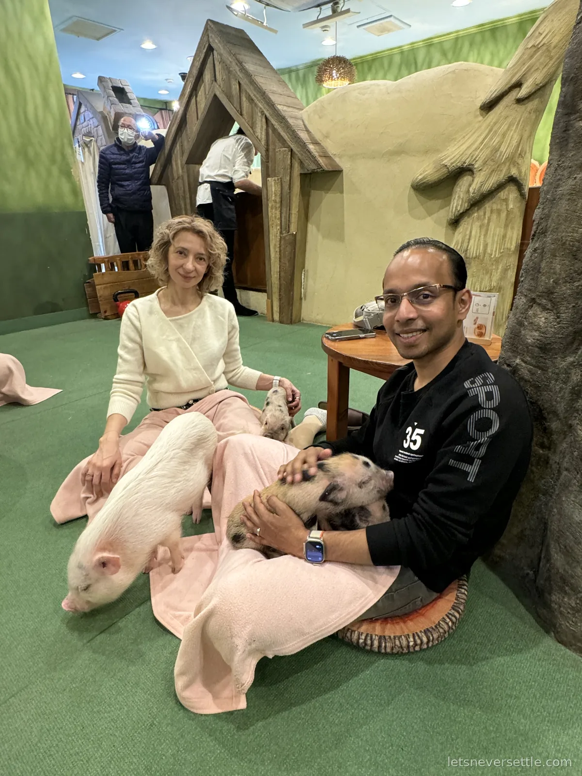 Inga and Dileep sitting on a green carpeted floor with pink blankets on their laps while micro pigs doze on them