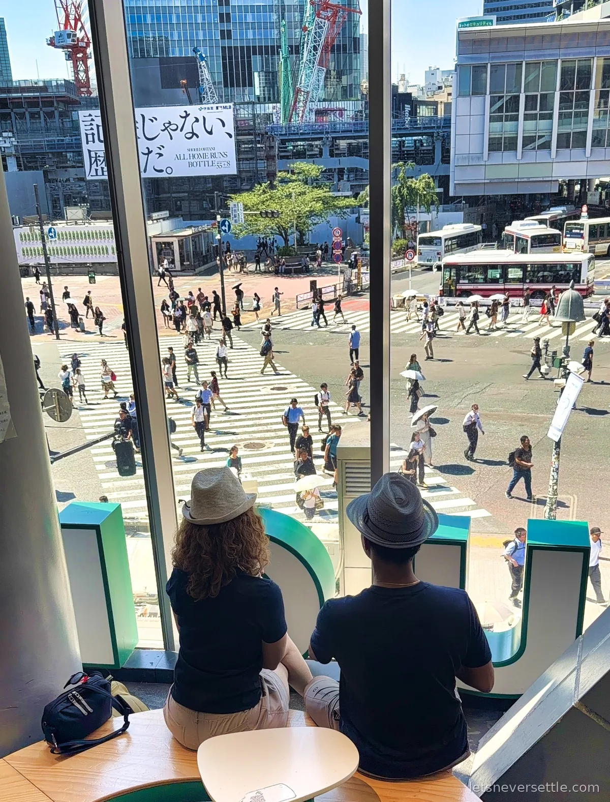 Inga and Dileep in sun hats seen from behind, watching Shibuya Crossing from a window seat above the intersection