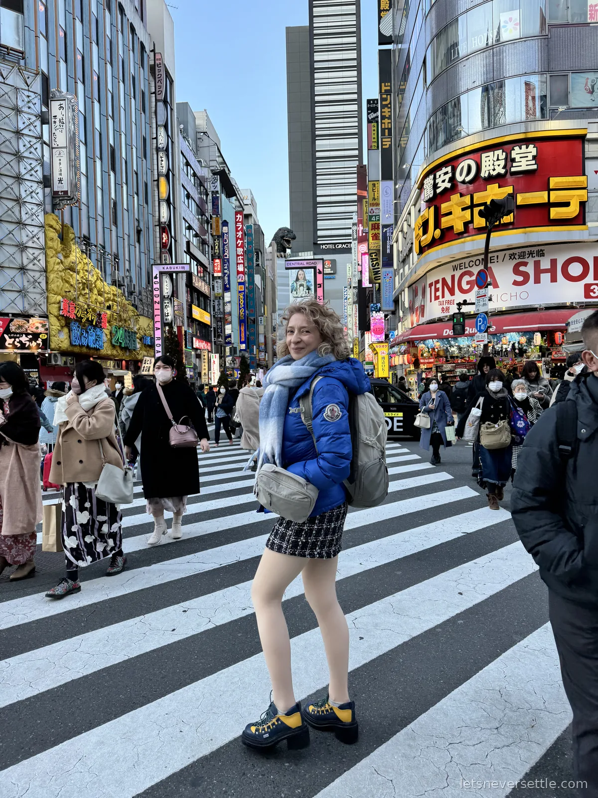 Inga in a blue puffer jacket standing on a zebra crossing in Shinjuku surrounded by shoppers and stacked neon signs