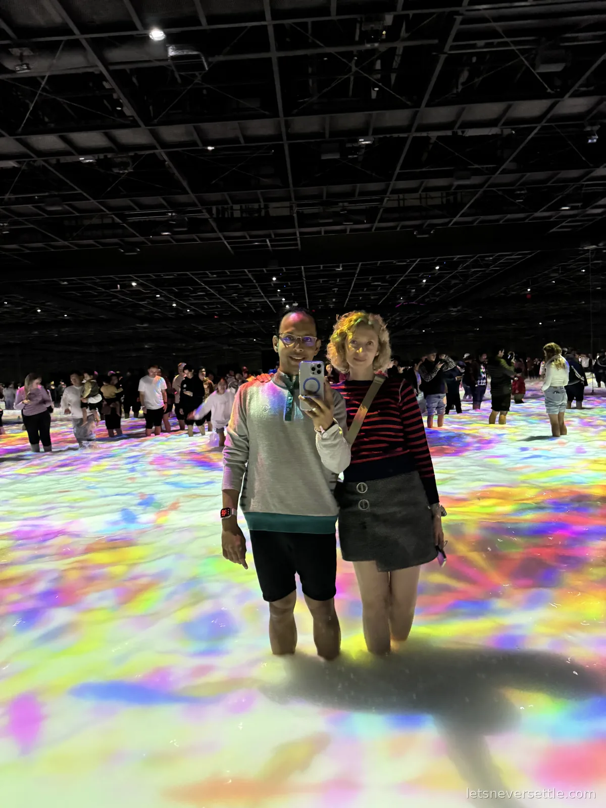 Inga and Dileep standing knee-deep in water covered with projected swirls of colour at the teamLab Planets digital art museum