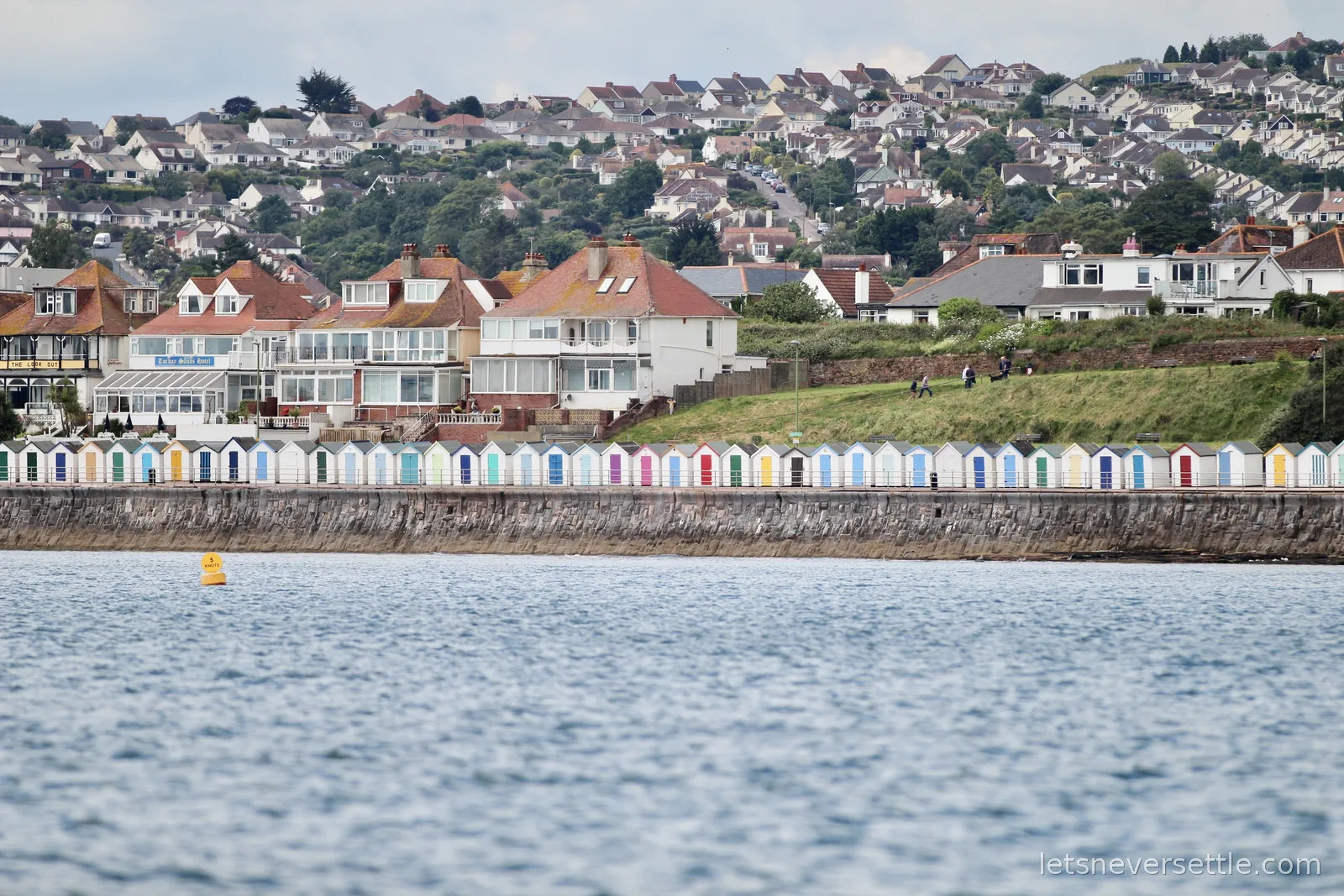 A long row of pastel-coloured beach huts along a sea wall with white seafront houses and a hillside of rooftops behind