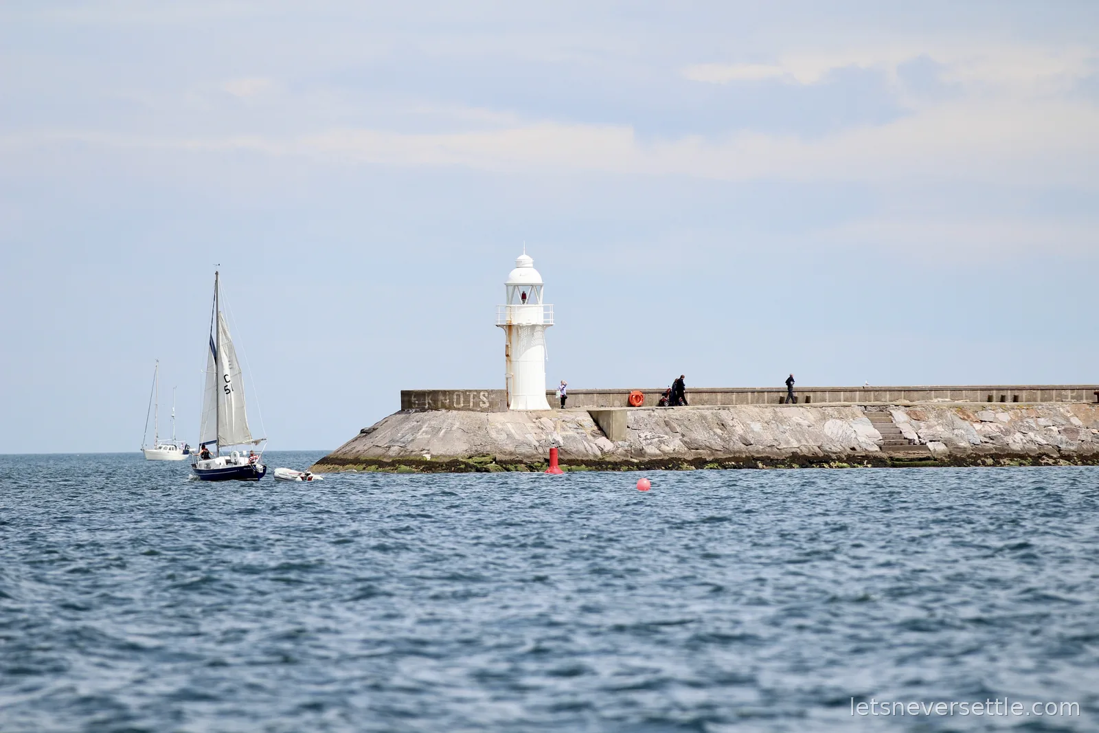 Small white lighthouse at the end of a stone breakwater with a sailing boat passing and figures walking on the pier